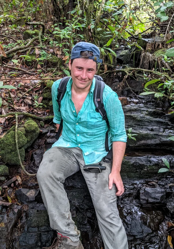 David Simons sitting on a rock in forest during fieldwork, with a backpack and a GPS unit.