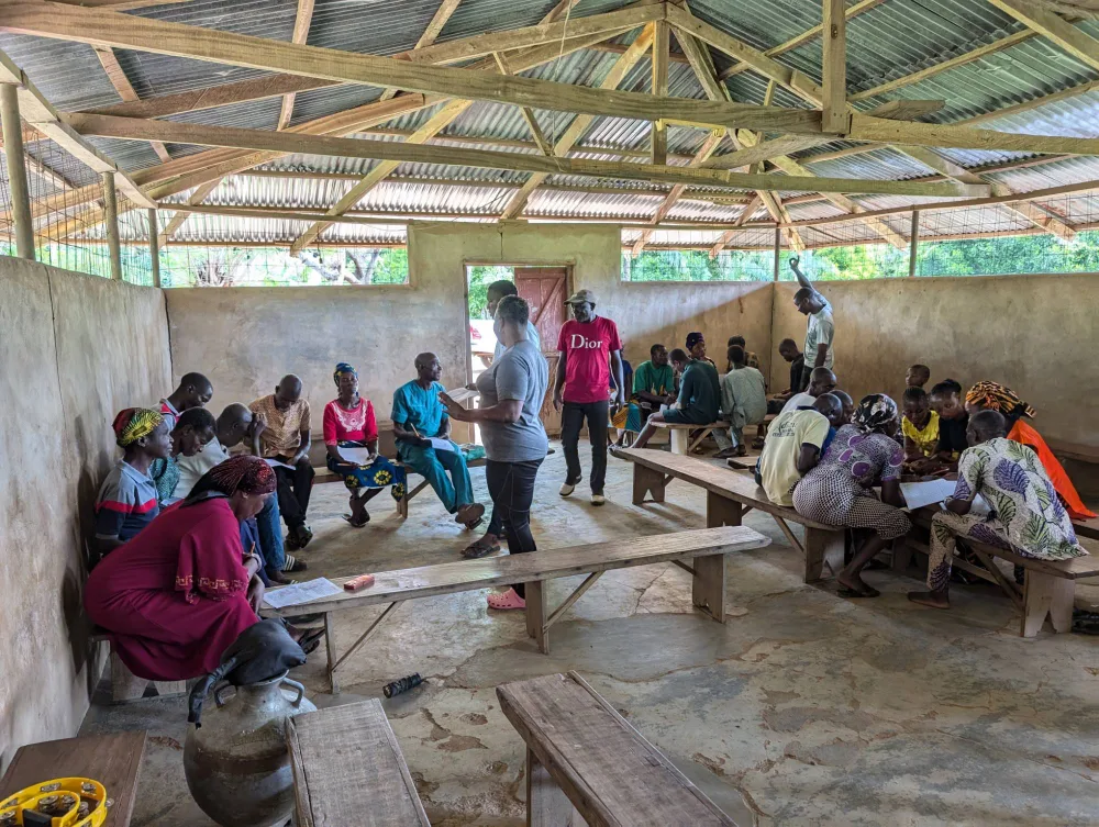 Community members seated on benches in a village hall, working through paper forms with members of the study team.